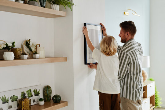 Back view portrait of father and son hanging picture together in minimal home interior decorated with succulents, copy space
