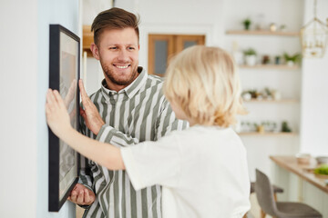 Fototapeta premium Side view portrait of happy father and son hanging picture together and looking at each other, copy space