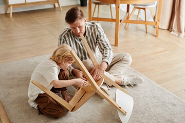 High angle portrait of father and son assembling wooden furniture together at home in earthy cozy...