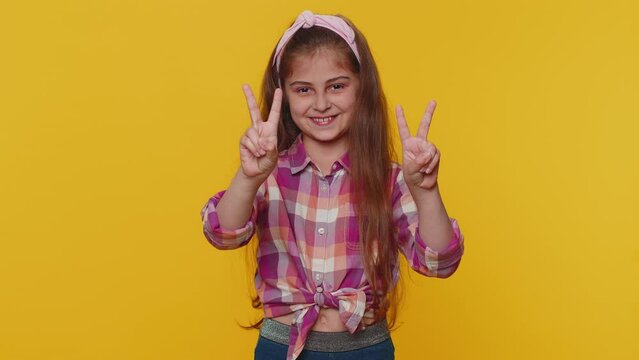 Happy preteen child girl kid showing victory sign, hoping for success and win, doing peace gesture smiling with kind optimistic expression. Little toddler children isolated on studio yellow background