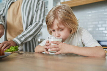 Portrait of cute blonde boy drinking milk in morning with father cooking in cozy kitchen interior, copy space