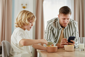 Side view portrait of father and son at breakfast in morning with dad using smartphone in background, copy space