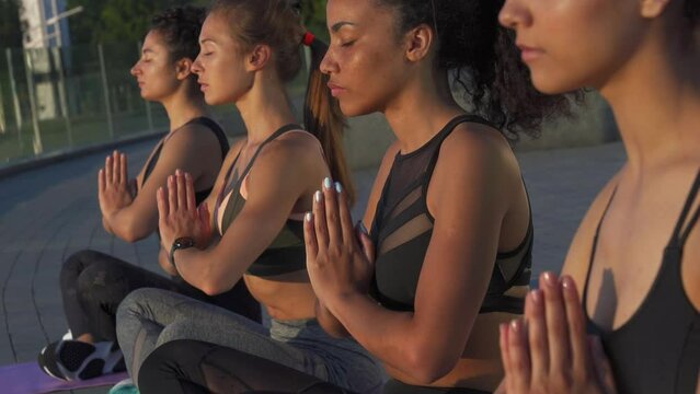Multiethnic young women meditate with yoga teacher in lotus pose breath fresh air at retreat yoga class on sunrise outdoor. Fit healthy multiracial ladies do yoga exercise in sunset on summer together