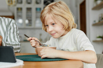 Portrait of young blonde boy using smartphone at dining table and watching videos online, copy space