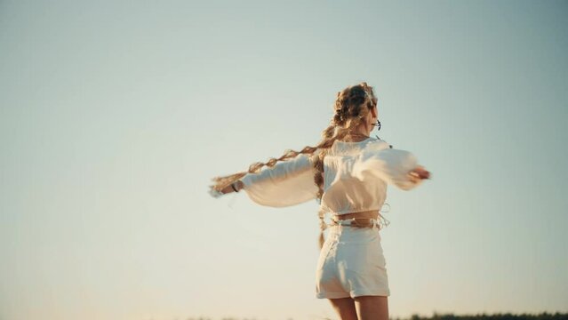 4K Slow-mo video. Happy young blond woman with braid spining on blue sky  background. Sunset cinematic consept. National ethnic costume. Freedom and happiness atmosphere.