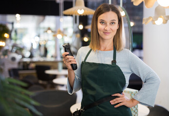 Portrait of smiling young female hairdresser standing in modern barbershop, holding electric hair clipper in hands - obrazy, fototapety, plakaty