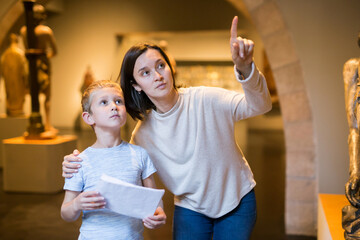 Young woman with son observing with interest sculptures exhibition in art museum, pointing to...