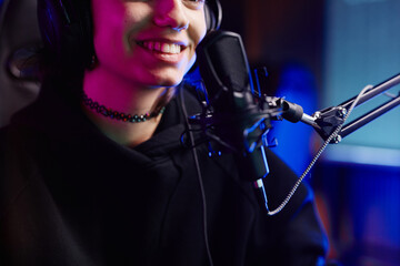 Close up of smiling young woman speaking to microphone in neon light while recording podcast or...