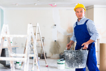 Man builder working in construction site, carrying bucket. Male repairer in overall and helmet lifting bucket.