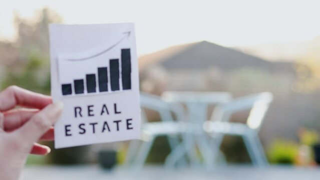 property value and return on investment or rent price, hand holding real estate sign with graph showing stats going up in front of backyard bokeh