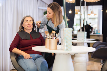 Young woman professional makeup artist working in beauty studio with positive elderly female client, applying tonal foundation on face.