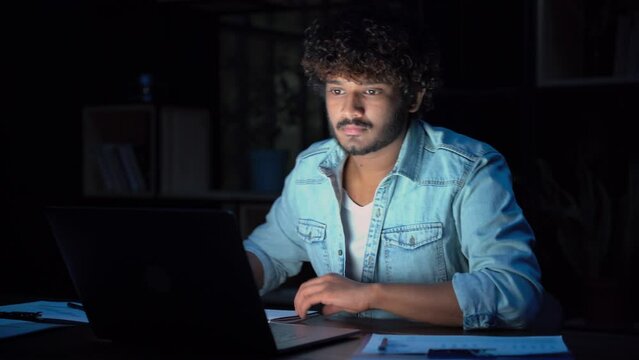 Young curly indian ethnic business man or student remote working overtime, learning online late at night at home or in dark office using laptop computer at workplace.