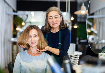 Pleasant young woman client sitting in chair while discussing haircut with professional elderly female hairdresser .