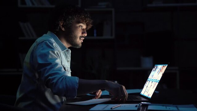 Young indian business man having remote video call conference team meeting working online by distant videocall web chat late at night at home in dark office using laptop computer.