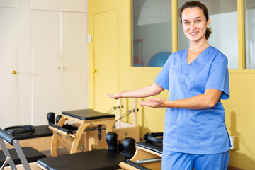 Smiling professional female physiotherapist polite inviting to rehab center with pilates equipment....