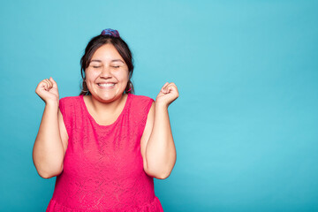 Cheerful curvy Latin woman with a wide smile and clenched fists, isolated on blue background.