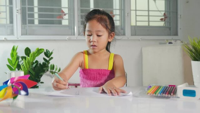 Child little girl drawing cartoon on paper before paint the color, Asian cute kid preschooler sitting on table smiling she draw and writing a picture with pencil at home to learning arts homework