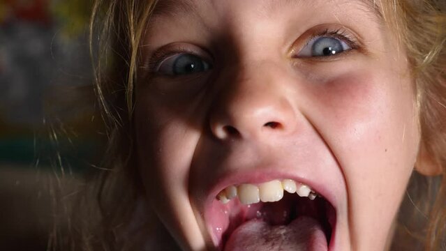 Blue-eyed girl firmly clenches her teeth, then opens her mouth wide, showing her tongue and soft palate. The tongue is far out. Close-up
