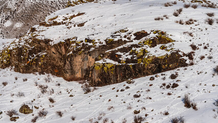 The red-brown rocks are covered with snow. Yellow and orange lichens are visible on the slopes. Dry grass and a scattering of stones are visible around. Altai