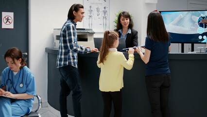 Mother and little girl writing checkup report at hospital reception desk to attend medical appointment. Patients having healthcare examination to receive support and assistance.