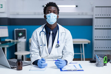 General practitioner in clinic cabinet sitting at desk while wearing facemask because of covid epidemic outbreak. Medic sitting in office while wearing professional uniform and protective gloves.