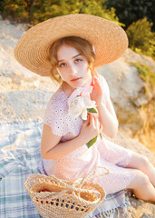 Elegant girl in a straw hat dressed in vintage style posing on a hill at sunset.