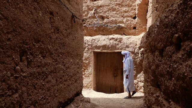 A traditional dressed Moroccan man wearing a white gandoura and a turban walks inside a Kasbah in Tamnougalt, Morocco. Authentic rural scene of South Morocco. 4k.
