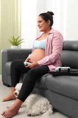Pregnant woman playing playstation sitting on couch, laptop, living room