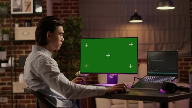 Asian businessman working on computer with greenscreen in living room. Looking at blank copyspace background with mockup template and isolated chroma key on display, modern technology.
