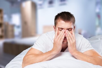Upset young man in pajamas waking up with headache, lying in bed early in morning, touching his...