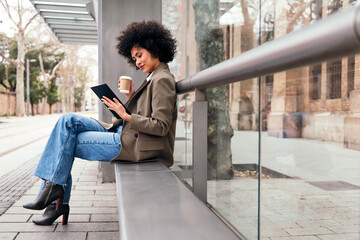 young latin woman sitting at the tram stop with a coffee in her hand and using a tablet, technology and urban lifestyle concept
