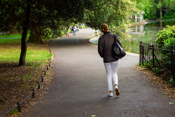 Slim woman in white jeans and black jacket walking in a park. Warm sunny day. Refreshing concept.
