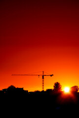 Tall crane silhouette against warm orange sunset sky. City skyline. Sun glow and flare. Dramatic rich saturated color.