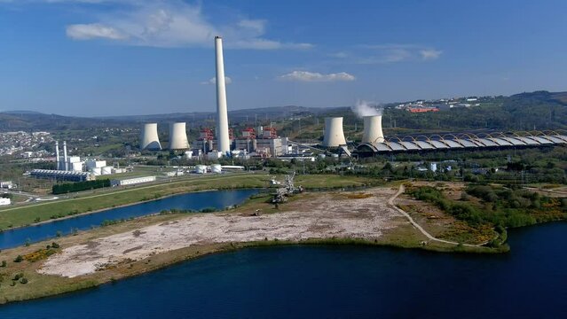 Steaming thermal power plant behind the lake with old excavator, a bridge on the right, sunny day. Drone shot traveling backwards very slowly. As Pontes, Lugo, Spain