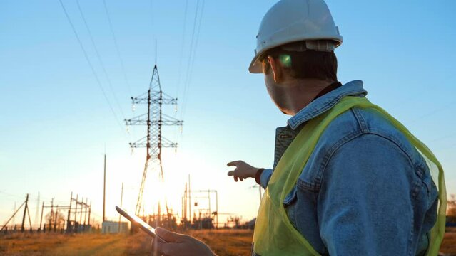Silhouette of engineer walk on field with electricity towers. Electrical engineer with high voltage electricity pylon at sunset background. Power workers at work concept.