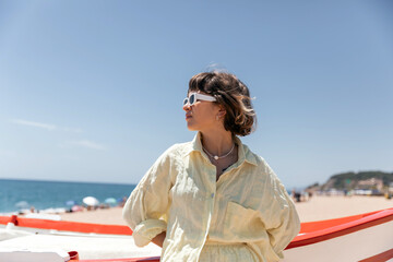Fototapeta na wymiar Caring pretty stylish woman with short hair wearing sunglasses and linen summer yellow suit is looking aside and resting on the beach on background of boat and blue sky
