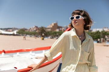 Adorable lovely charming girl in summer stylish outfit and sunglasses is posing in sunny warm day on the beach near the boat. Concept of enjoying weekend, vacation