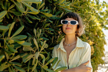 Pretty smiling lovely woman with wavy short hair in sunglasses wearing yellow summer shirt posing on green plants background in summer sunny day