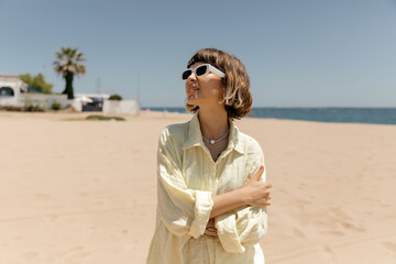 Lovely stylish european girl with short wavy hair wearing linen yellow shirt in sunglasses is looking aside with smile and resting on sandy beach in summer sunny day