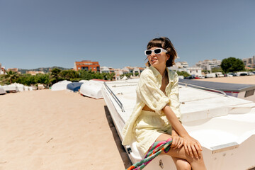 Adorable lovely smiling girl with wavy short hair in white sunglasses wearing yellow summer suit is sitting on the boat on sandy beach in summer sunny day against blue sky