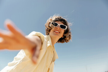 Pretty charming young girl with wavy short hair in sunglasses wearing summer yellow shirt waving at...