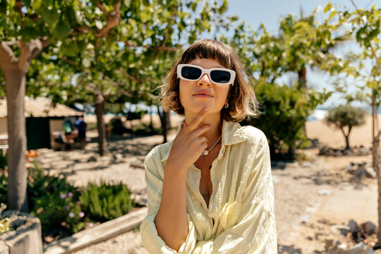 Young caucasian beautiful lady with short hair wearing sunglasses and yellow linen shirt while standing on beach with green plants background. Brown-haired woman in sunglasses 