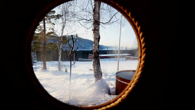 View from behind the round window of the bathhouse to the font with hot water outside in winter at low sub-zero temperatures