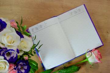 Flatlay. Notepad, diary with a pen on a wooden table near a bouquet of multi-colored roses. Workspace with diary and pink white flower on wooden background. Top view