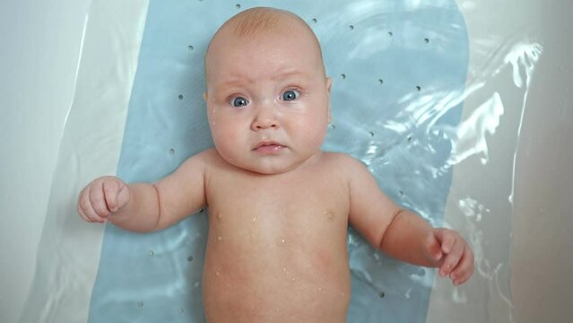 Baby girl lies in bathtub with water. Child looks around. Baby daughter likes to bathe waving hands with wonderment closeup