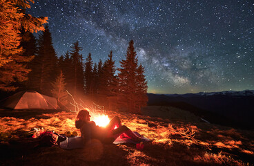 Female hiker lying at karemat near bonfire admire beauty of stunning starry sky during night in campsite, backpacker visiting mountains with tent near spruce forest in National Park - obrazy, fototapety, plakaty