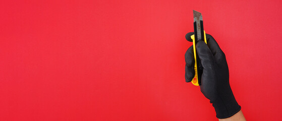 Hand wearing black gloves holding yellow cutter knife isolated red background