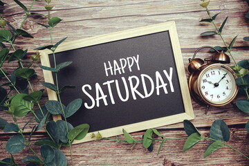 Happy Saturday typography text written on wooden blackboard with alarm clock and green eucalyptus...