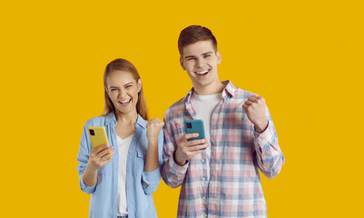 Overjoyed young friends Caucasian man and woman experience positive emotions when they see notification on mobile phone about great shopping offer dressed in casual clothes, stand in yellow studio