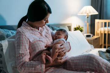 loving asian mother sitting on bed in pajamas is putting her cute baby in her cuddle to sleep at a cozy bedroom during nighttime at home.